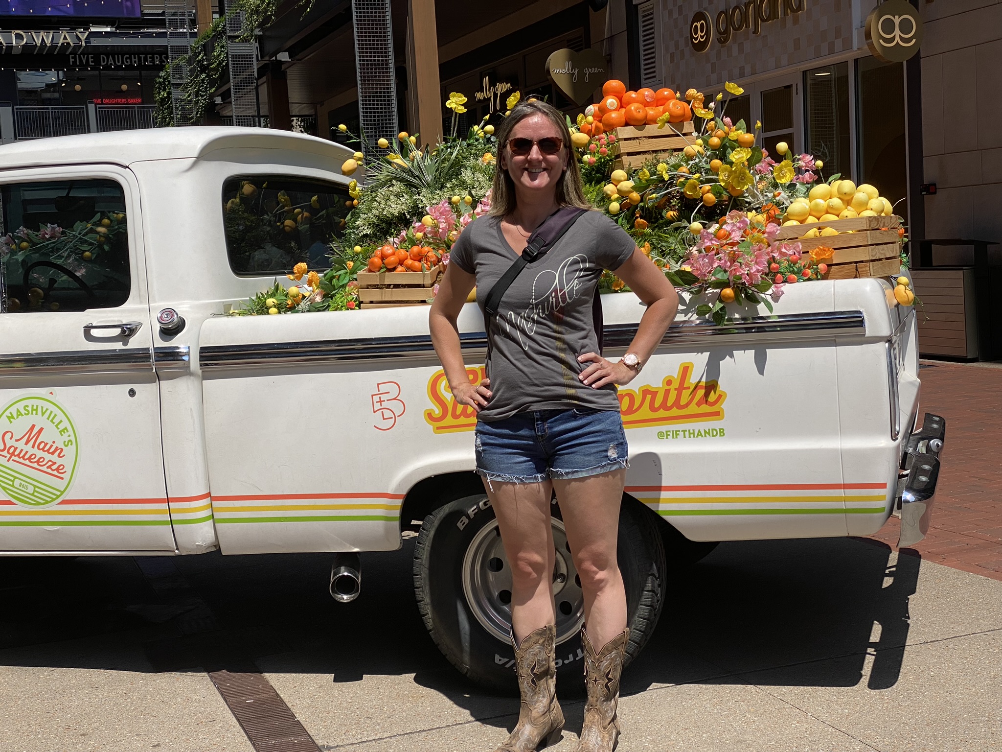 Kat, a white woman with short blonde hair, stands with her hands on her hip in front of an old white truck with orange and yellow fruit and flowers in the truck bed. She's wearing a grey v-neck Nashville t-shirt and short denim shorts and brown cowboy boots, as well as sunglasses. She's smiling.