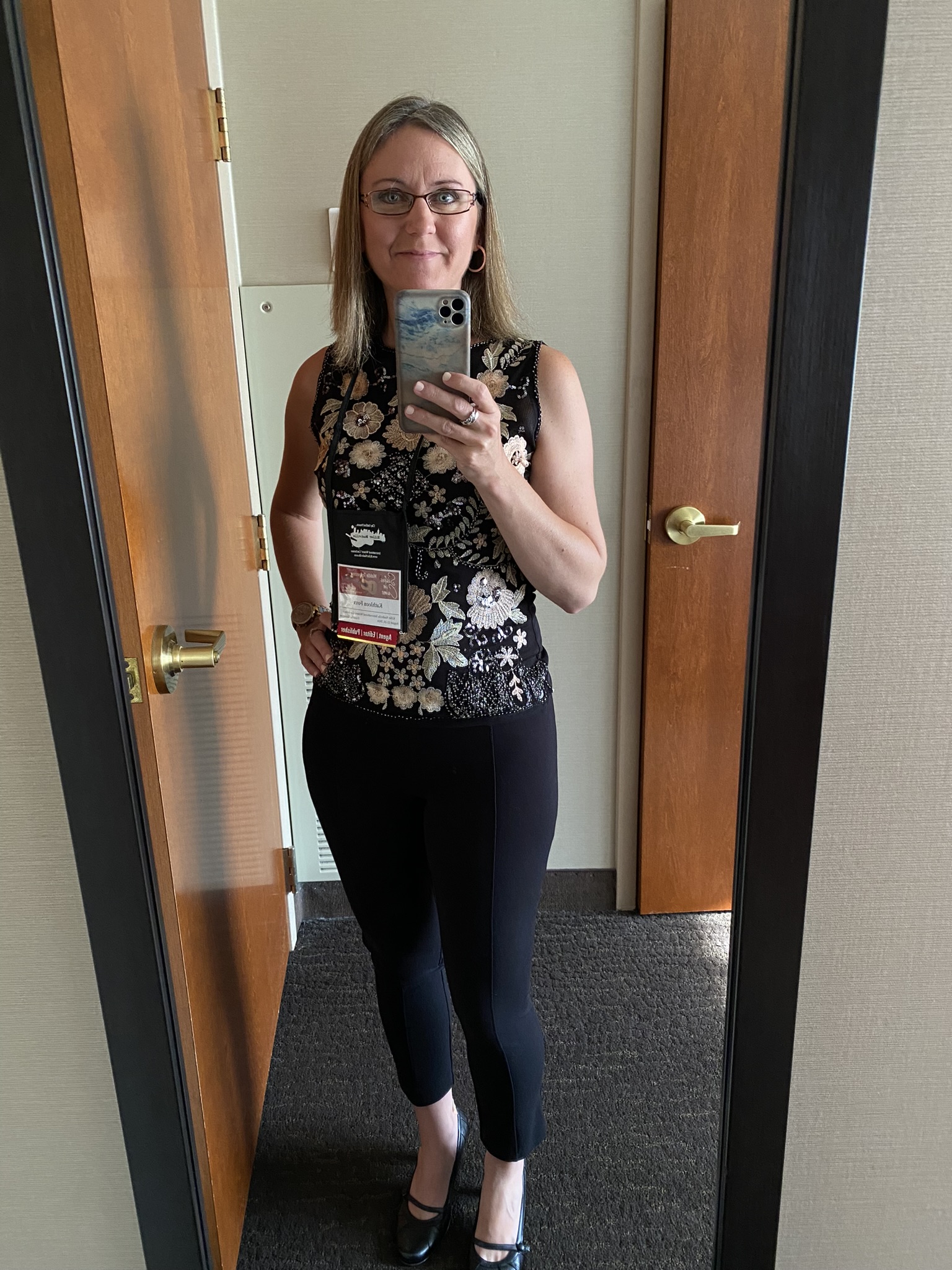 Kat standing in front of a mirror wearing black dress pants and a beautiful sequined sleeveless top with flowers on the front